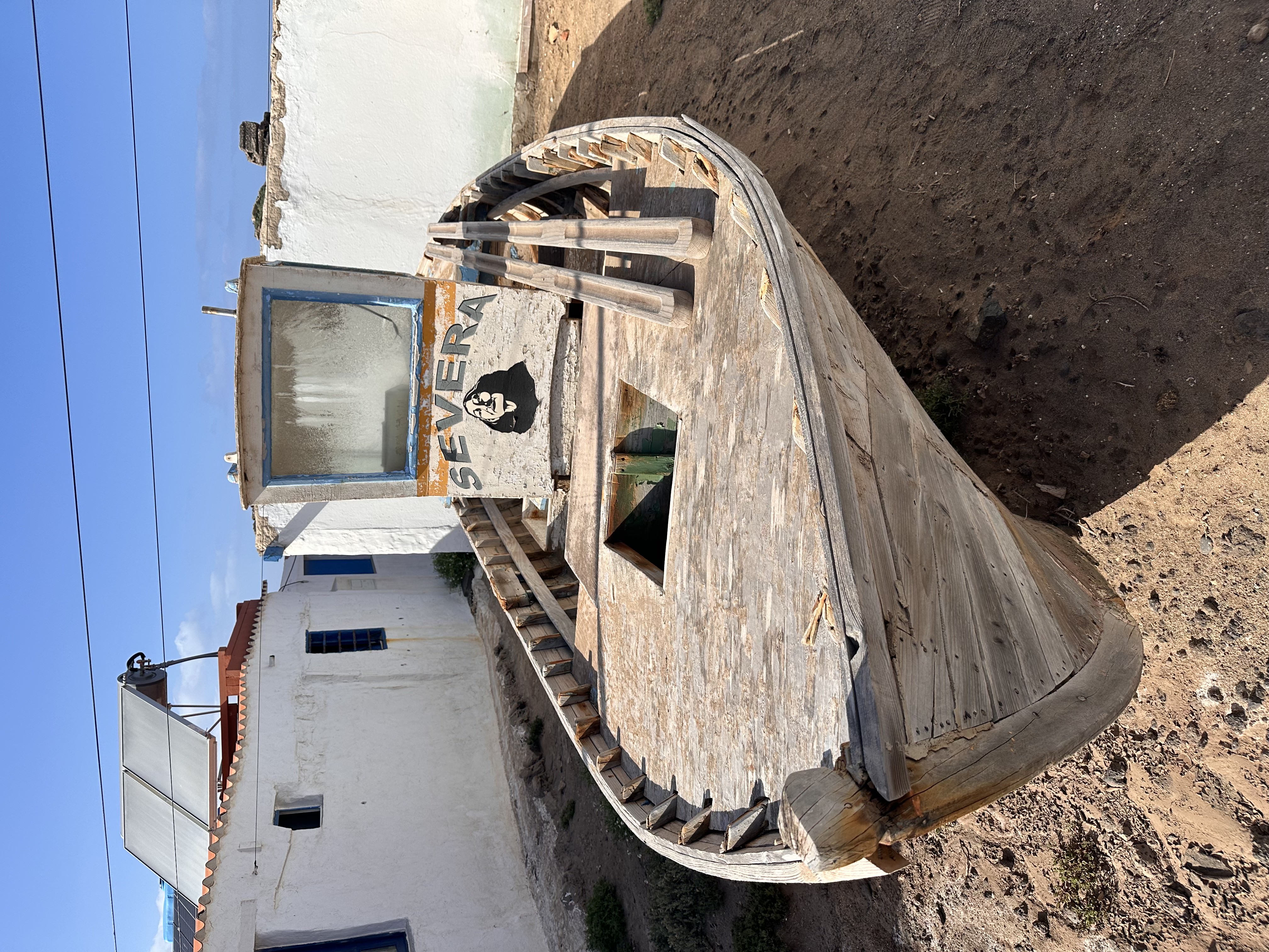 An old weathered wooden fishing boat named 'Severa', dry-docked between whitewashed houses with blue-framed windows and solar panels