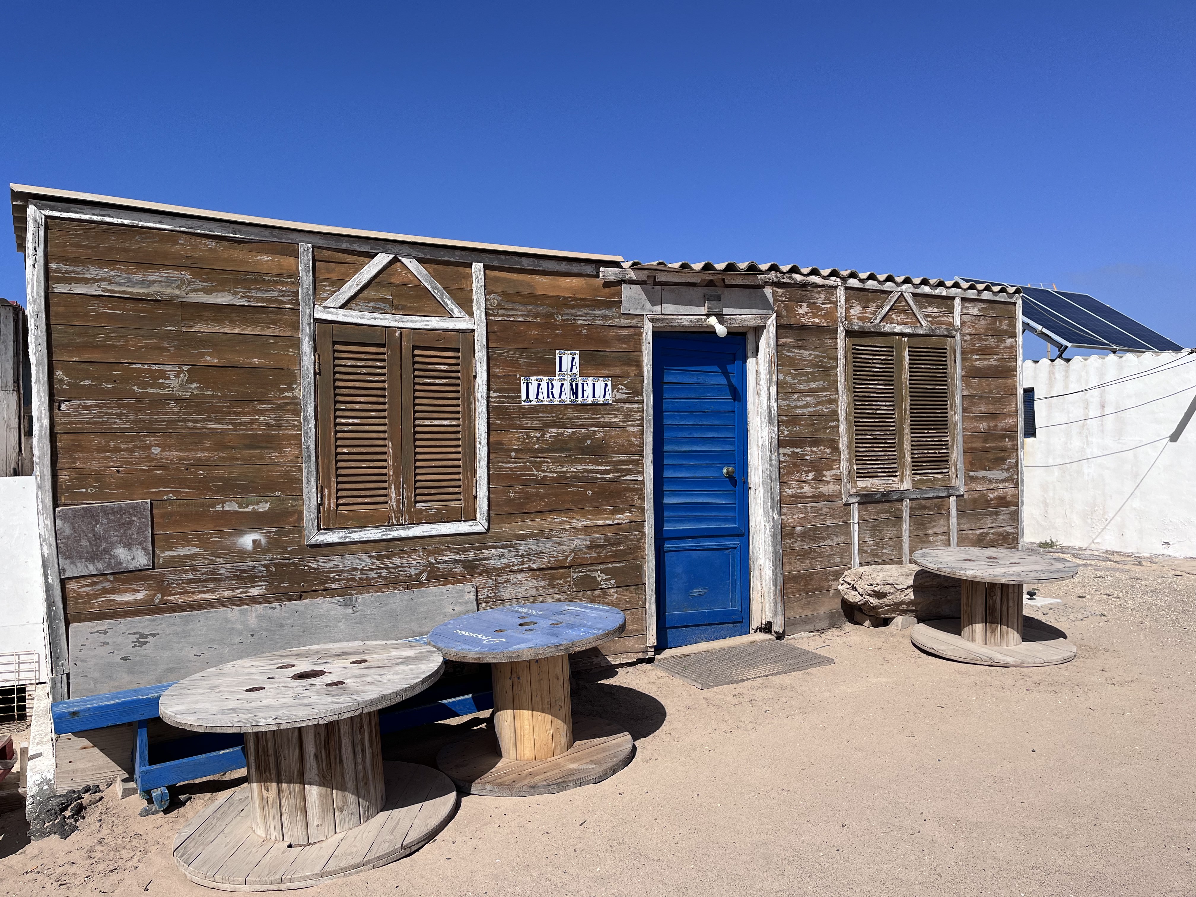 La Taramela — a rustic wooden shack with a vivid blue door, cable-drum tables out front, solar panels on the roof