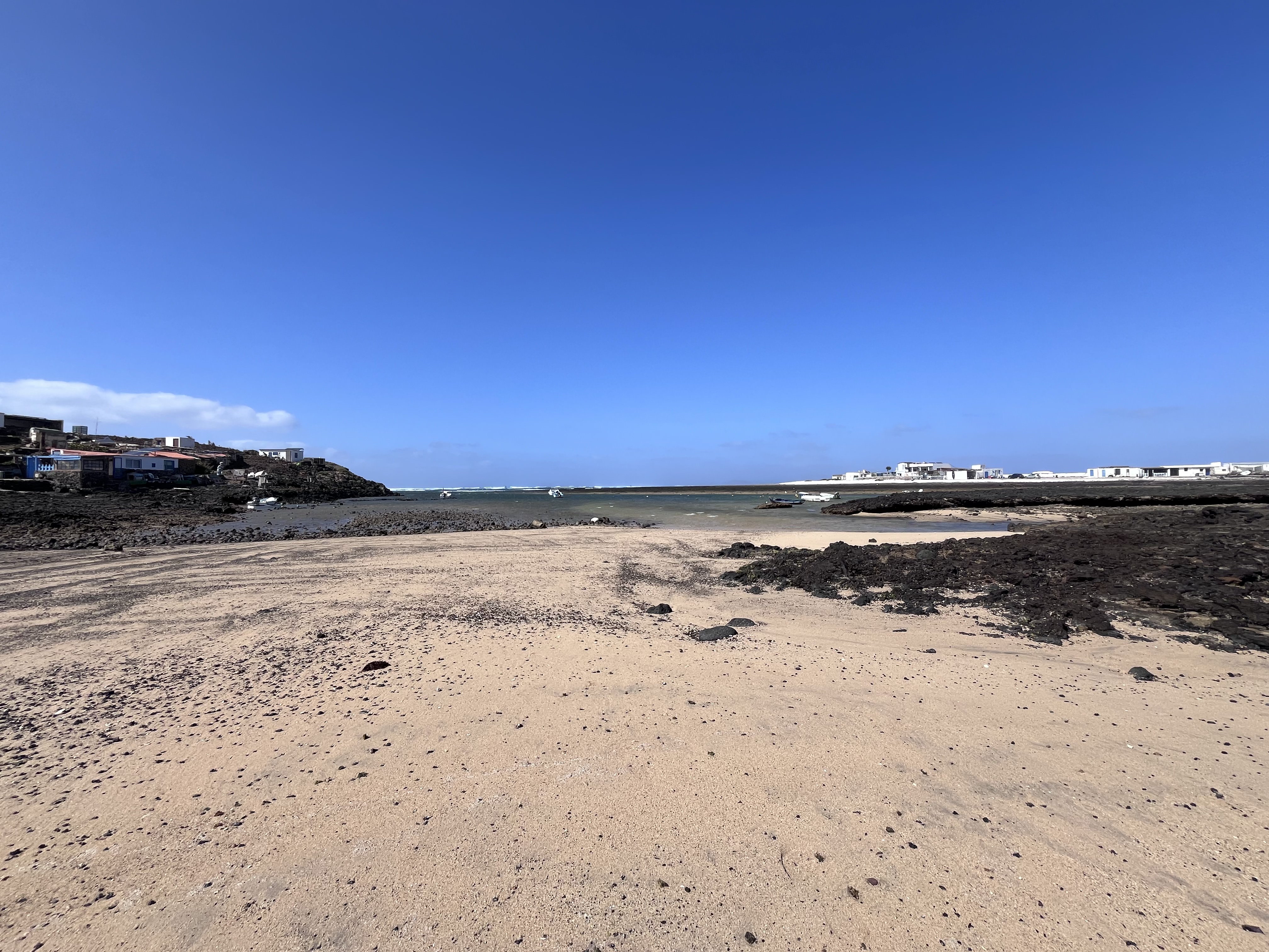 Wide view of Majanicho bay — sand, black volcanic rock, scattered boats, and the cluster of white buildings on each headland under a deep blue sky