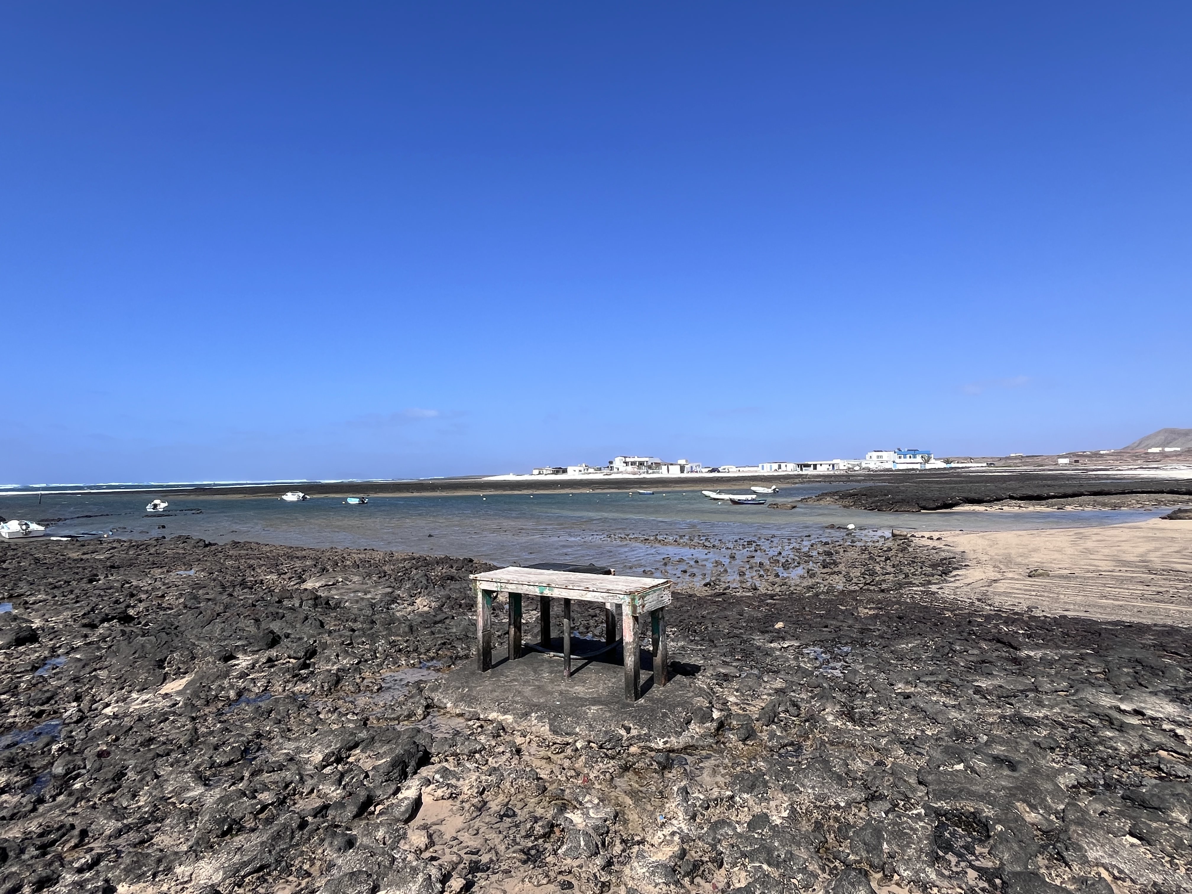 The traditional fish-cleaning table standing alone on the volcanic rock shelf at the edge of the bay, small boats and the village behind
