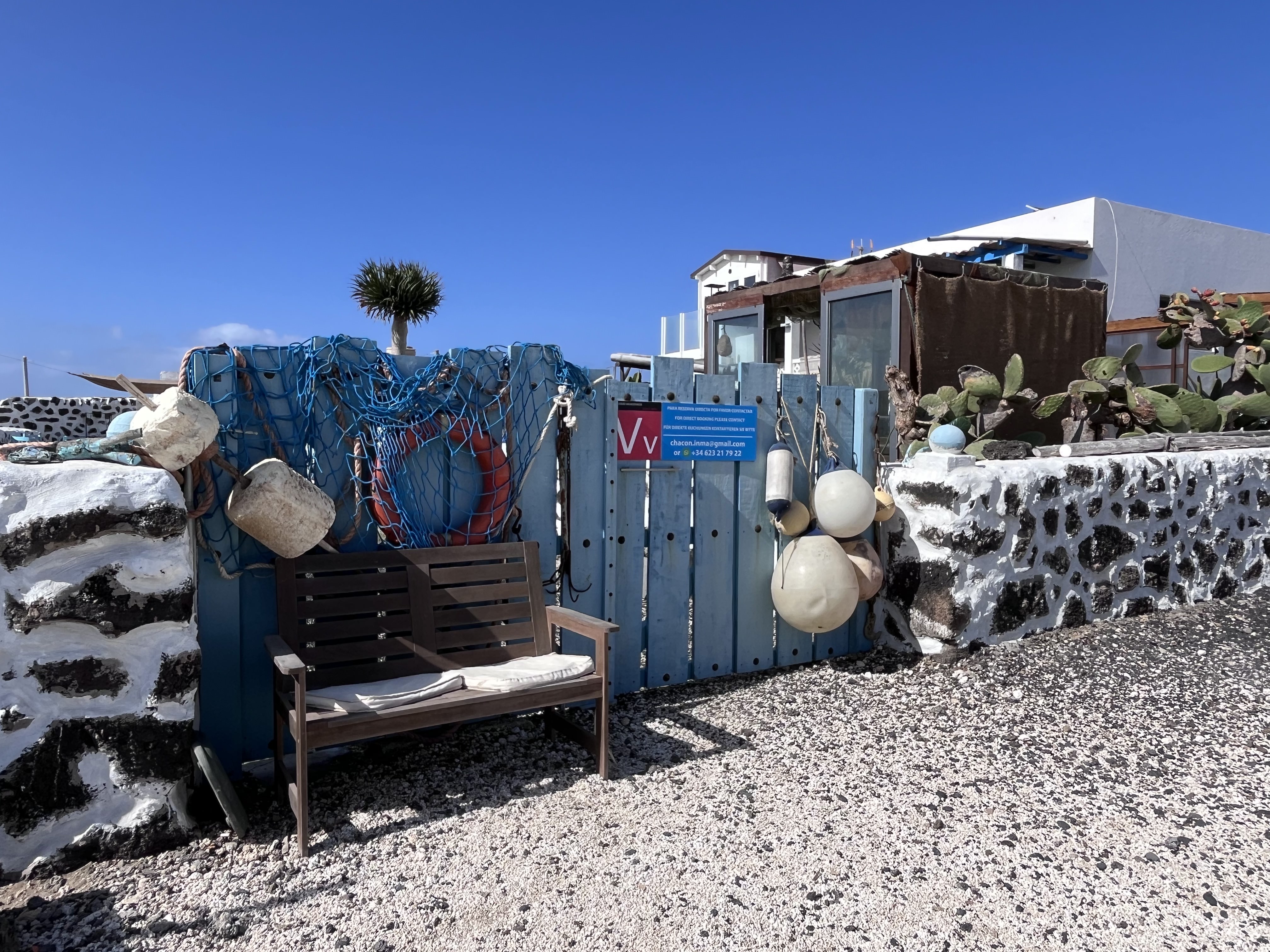 A gate in Majanicho decorated with blue fishing nets, rope buoys, and white floats — a bench sits outside in the Canarian sun