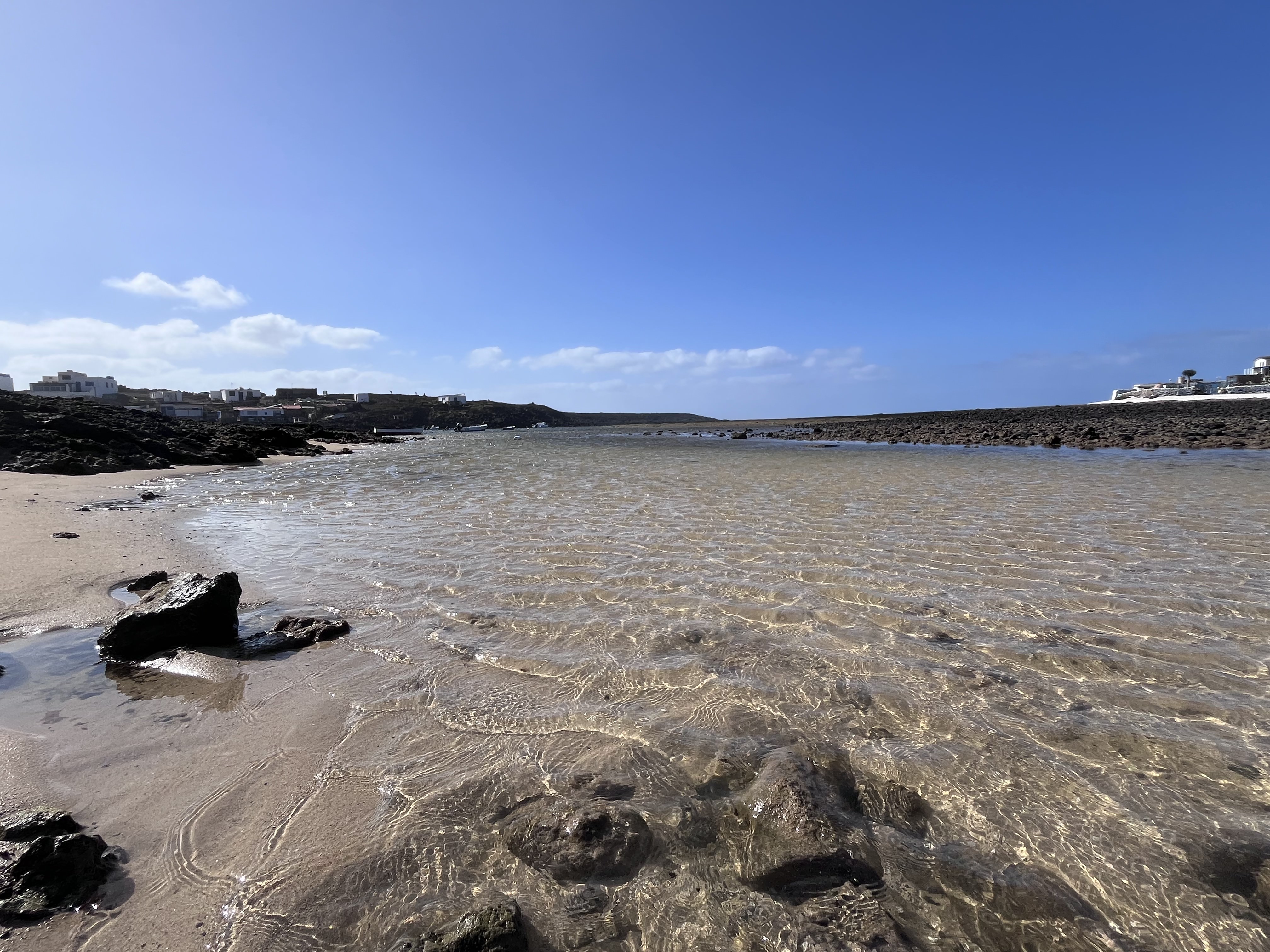 Crystal-clear, perfectly still shallow water in the bay at Majanicho — transparent over sand and volcanic rock, the village visible on the headland behind